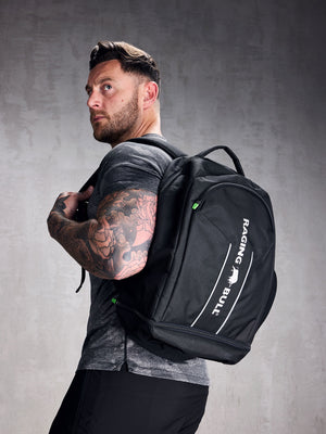 A man wearing a black rucksack with adjustable shoulder straps and a padded mesh back. The backpack features multiple compartments, including a zipped front compartment and a shoe compartment.