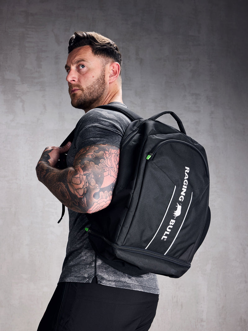 A man wearing a black rucksack with adjustable shoulder straps and a padded mesh back. The backpack features multiple compartments, including a zipped front compartment and a shoe compartment.