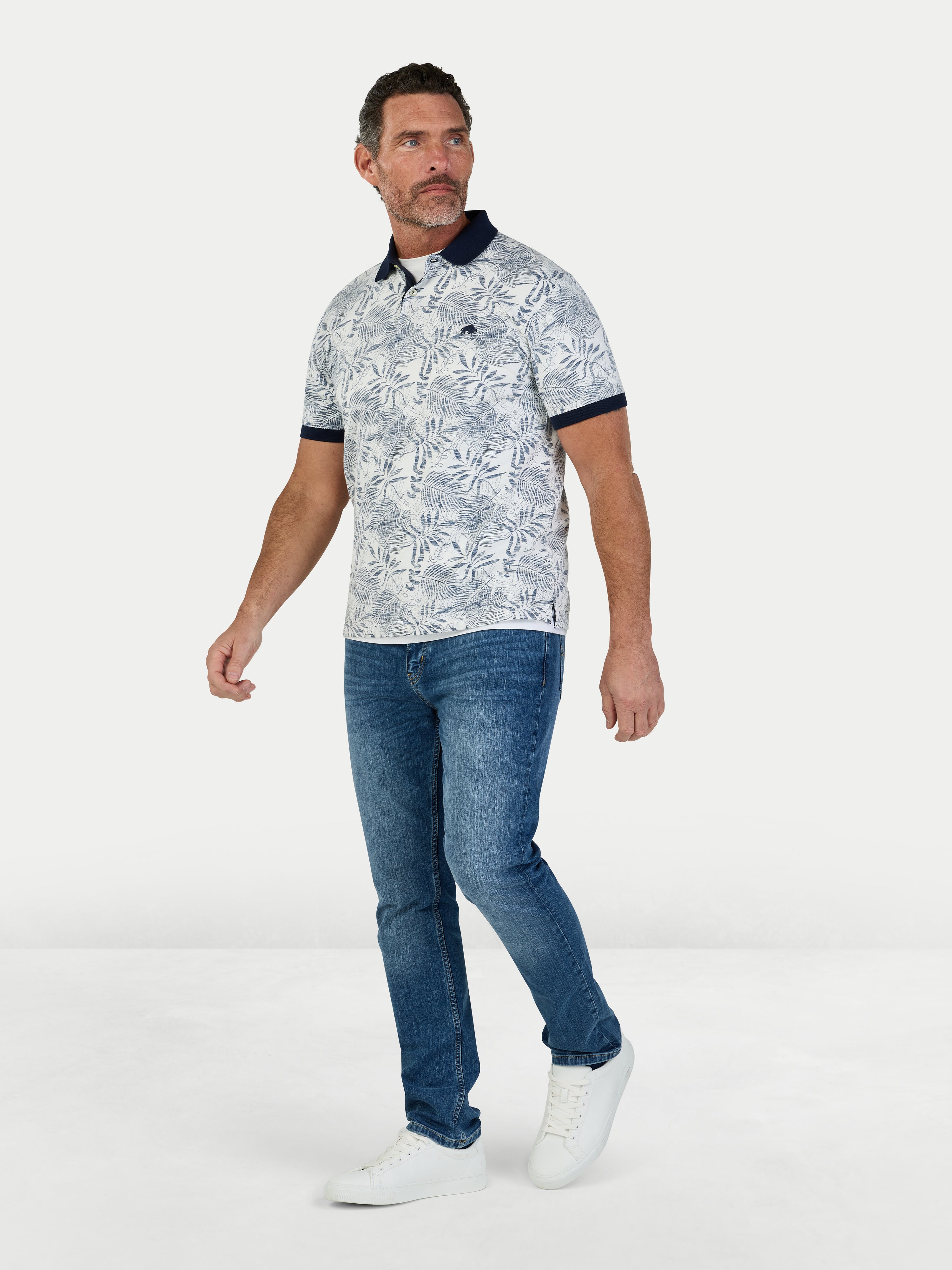 Man wearing a patterned polo shirt and blue jeans on a white background