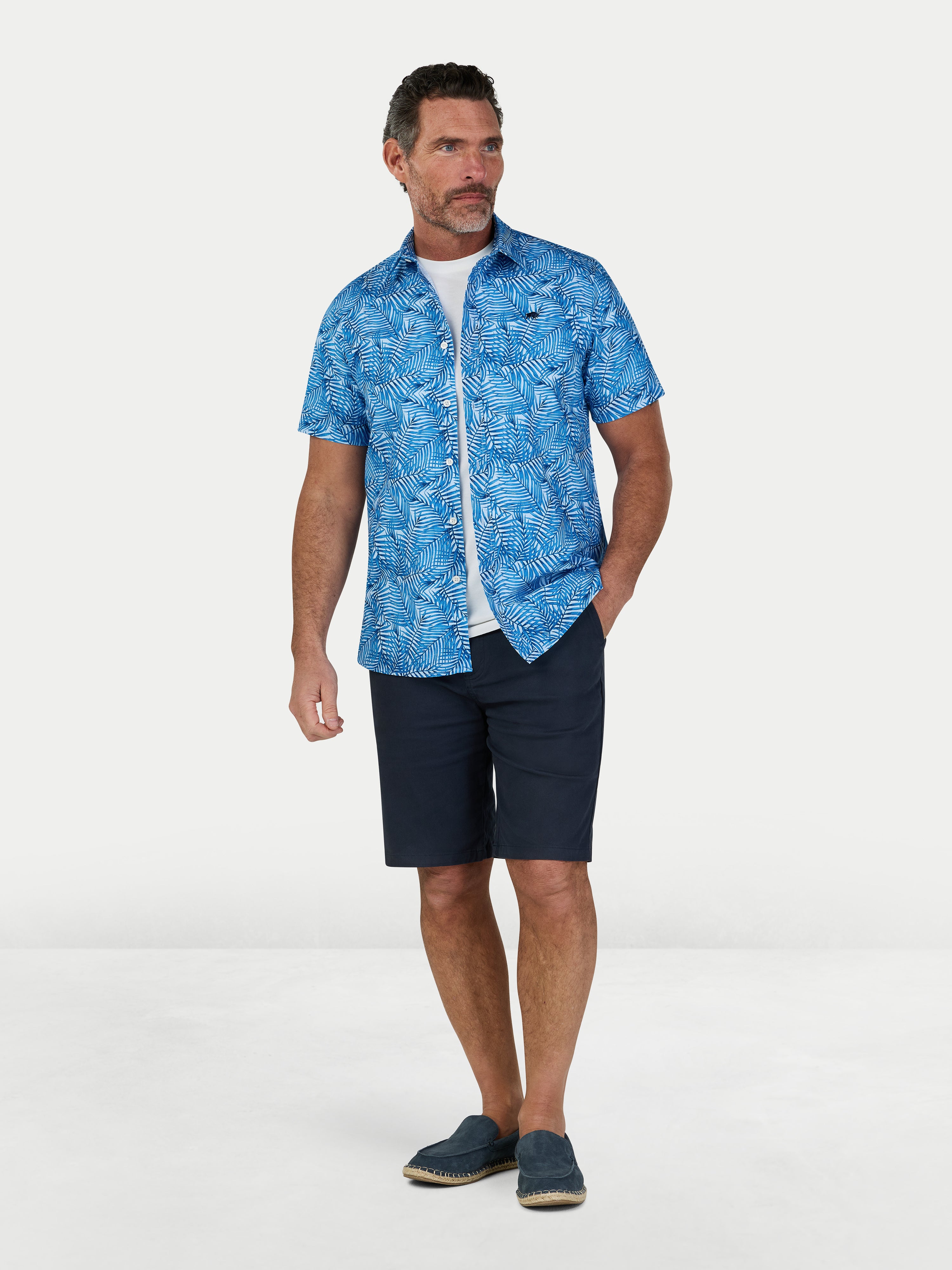 Man wearing a blue patterned shirt and navy shorts on a white background