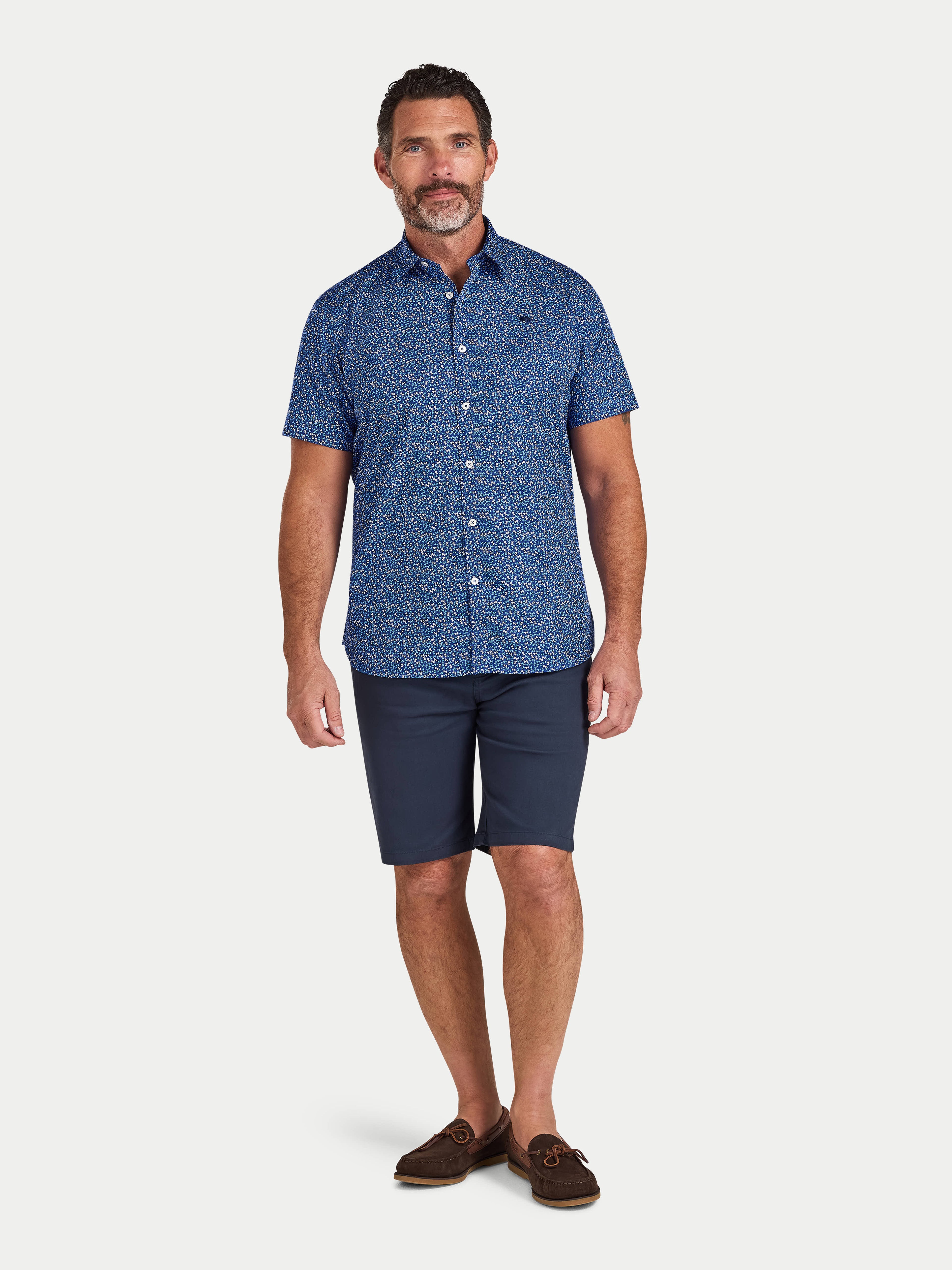 Man wearing a blue patterned shirt and navy shorts on a white background