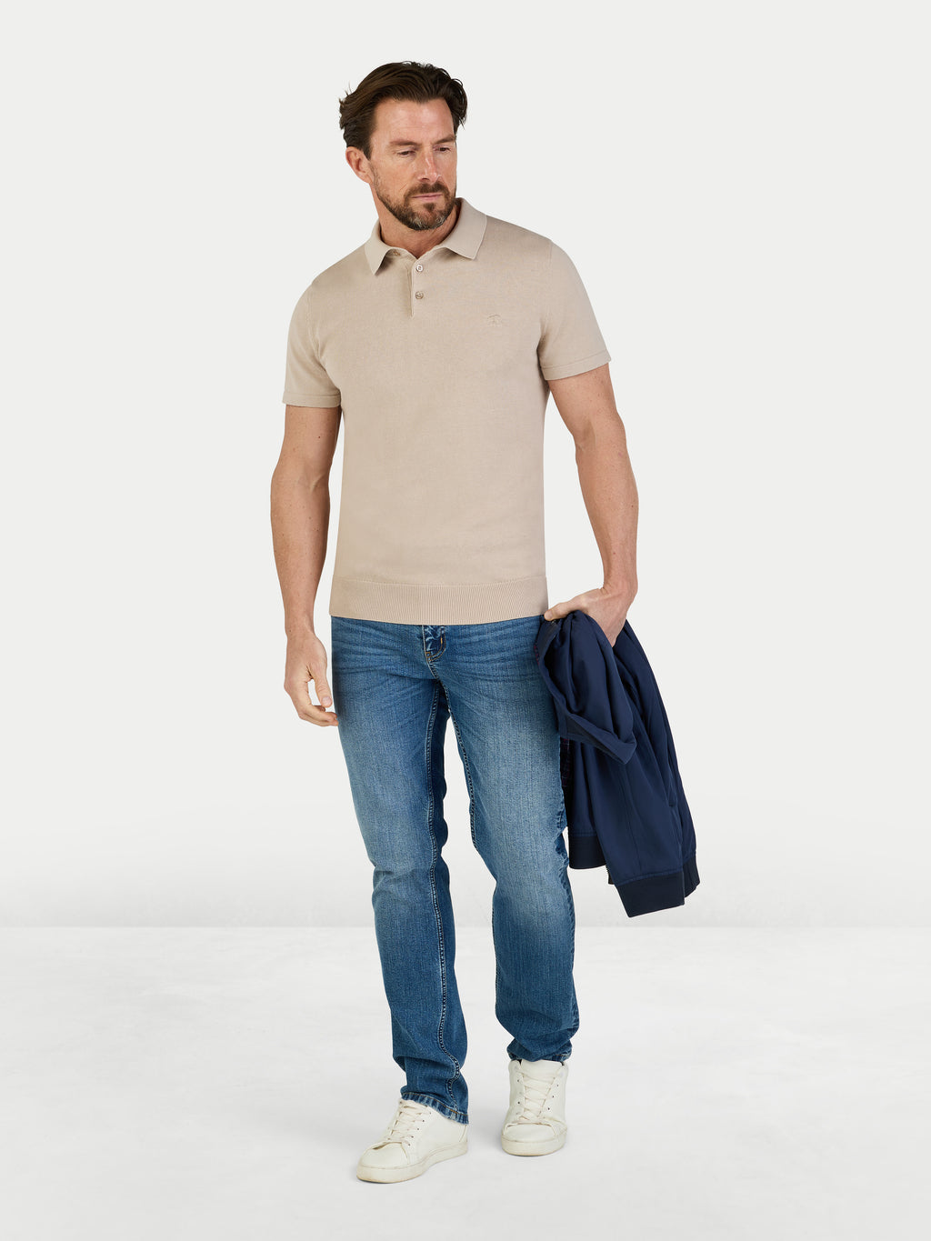 Man wearing a beige polo shirt and blue jeans holding a navy jacket on a white background