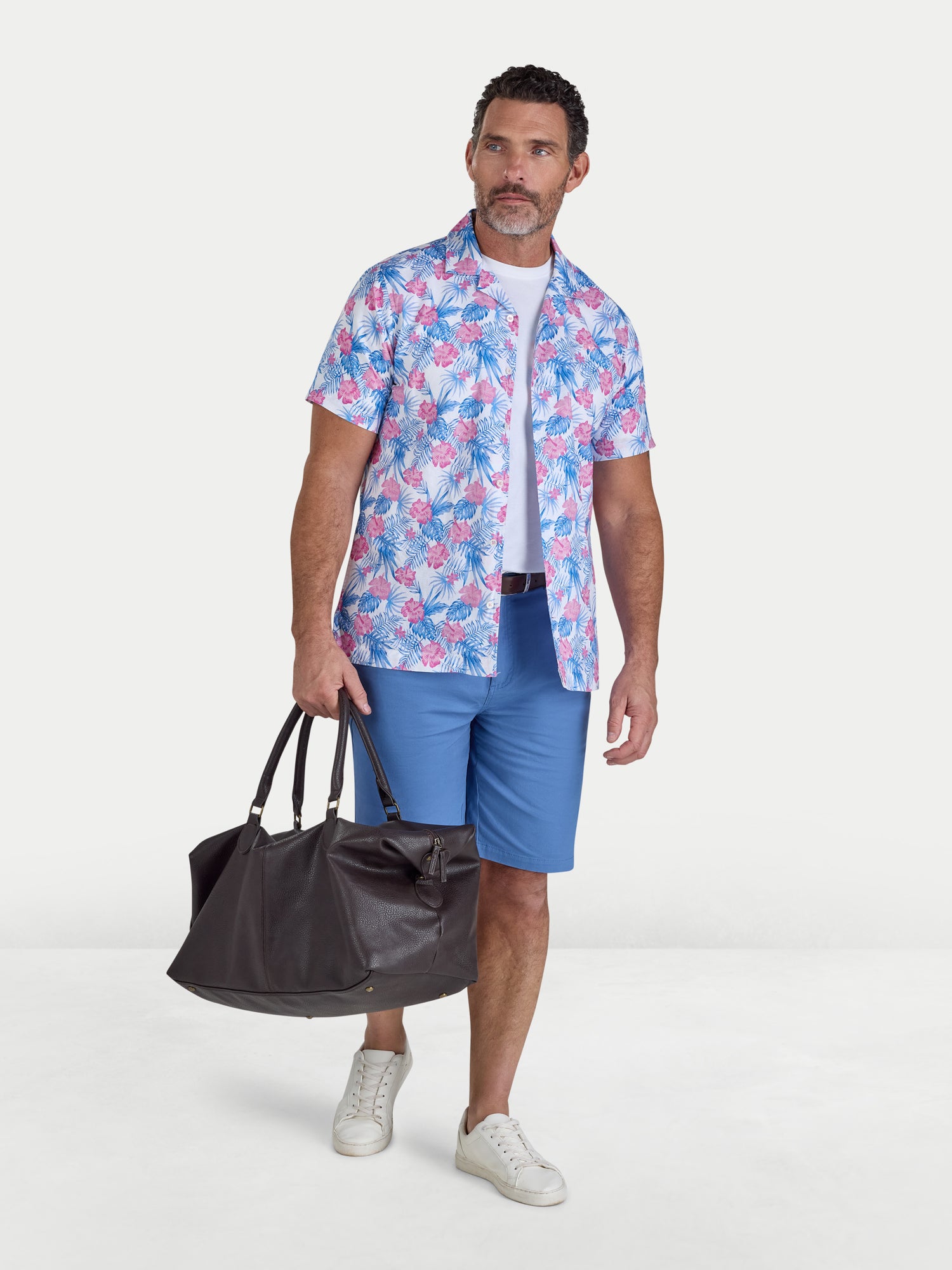 Man wearing a colorful floral shirt and blue shorts, holding a black duffel bag on a white background