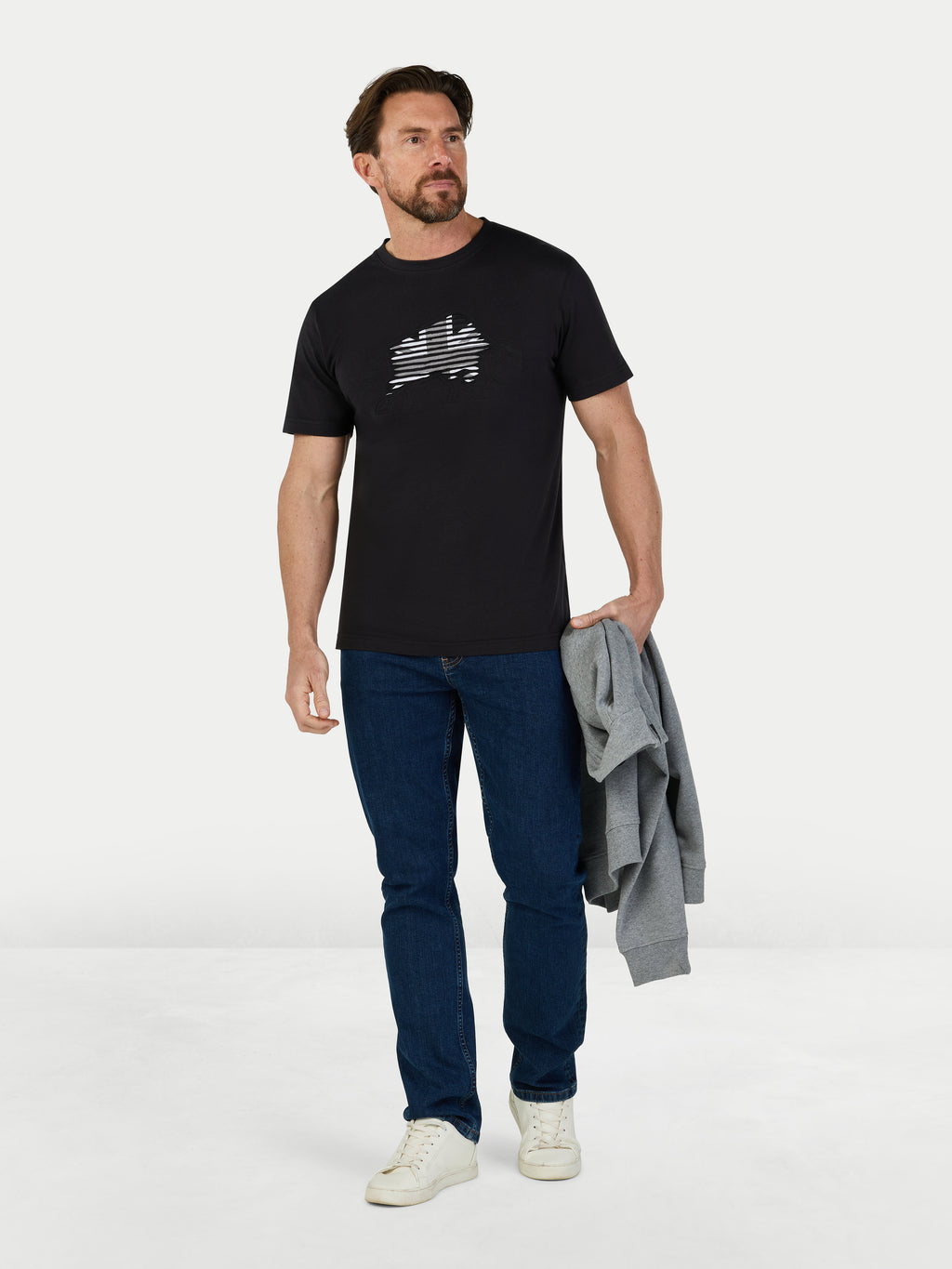 Man wearing a black t-shirt with a logo, blue jeans, and white sneakers on a white background
