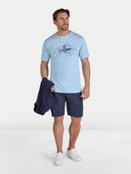 Man wearing a light blue t-shirt with a graphic design and dark shorts, holding a navy jacket on a white background.