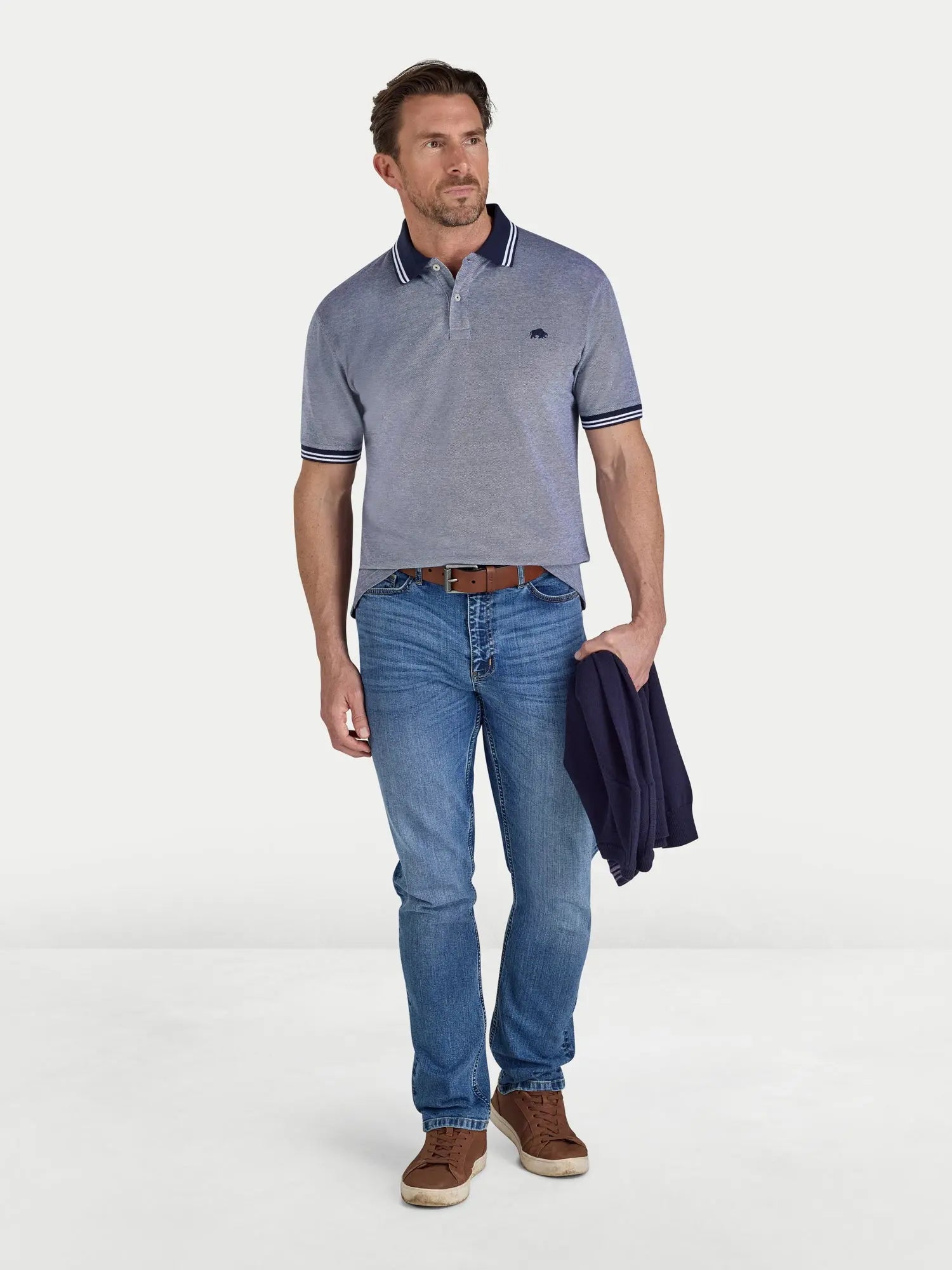 Man wearing a blue polo shirt and jeans holding a jacket on a white background